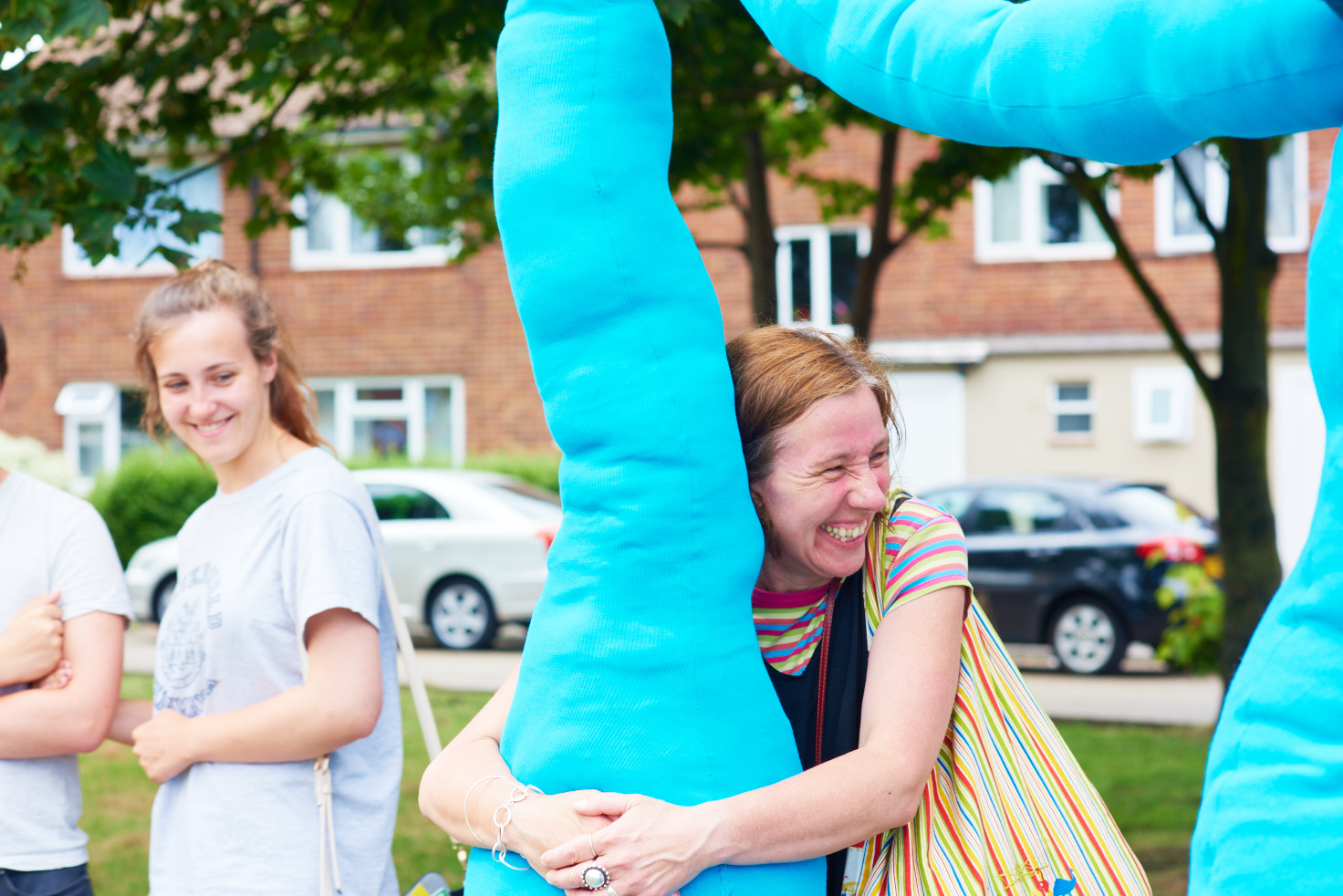 A photo showing an art installation on a green in front of a row of houses. The installation is large blue fabric tubes strung up about 2.5 metres in the air, with sections hanging down into large lumpy bulbs just off the ground. A woman is hugging one of the bulbs and laughing. Someone behind her is watching and smiling.