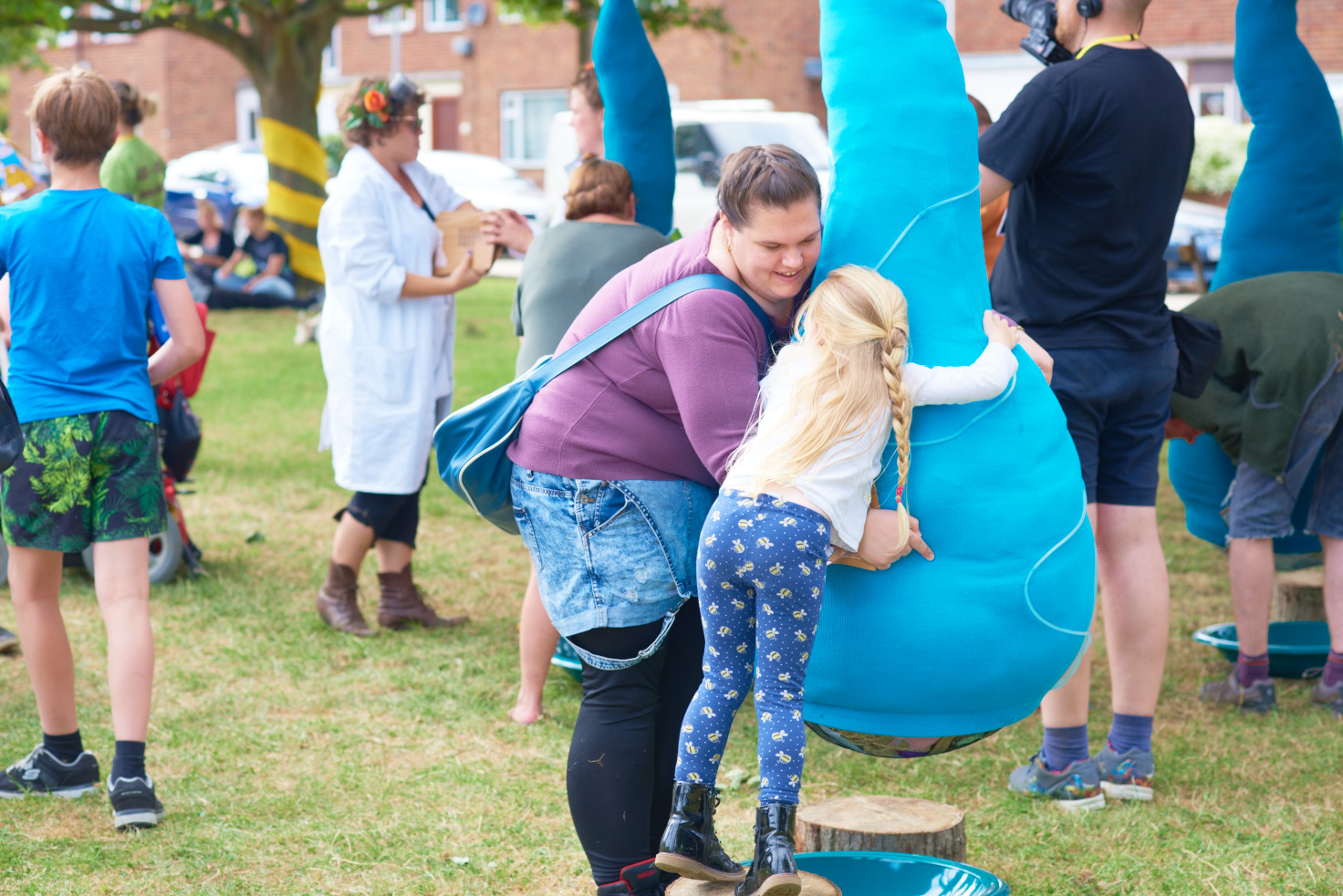 A photo showing an art installation on a green in front of a row of houses. The installation is large blue fabric tubes strung up about 2.5 metres in the air, with sections hanging down into large lumpy bulbs just off the ground. A woman and child are hugging the nearest bulb, the girl is standing on a circle of tree trunk about 15cm high in order to hug the bulb.