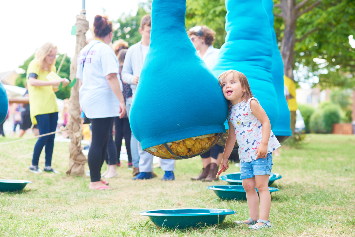 A photo showing an art installation on a green in front of a row of houses. The installation is large blue fabric tubes strung up about 2.5 metres in the air, with sections hanging down into large lumpy bulbs just off the ground.A young child is standing next to one of the bulbs with her head pressed against the blue fabric.