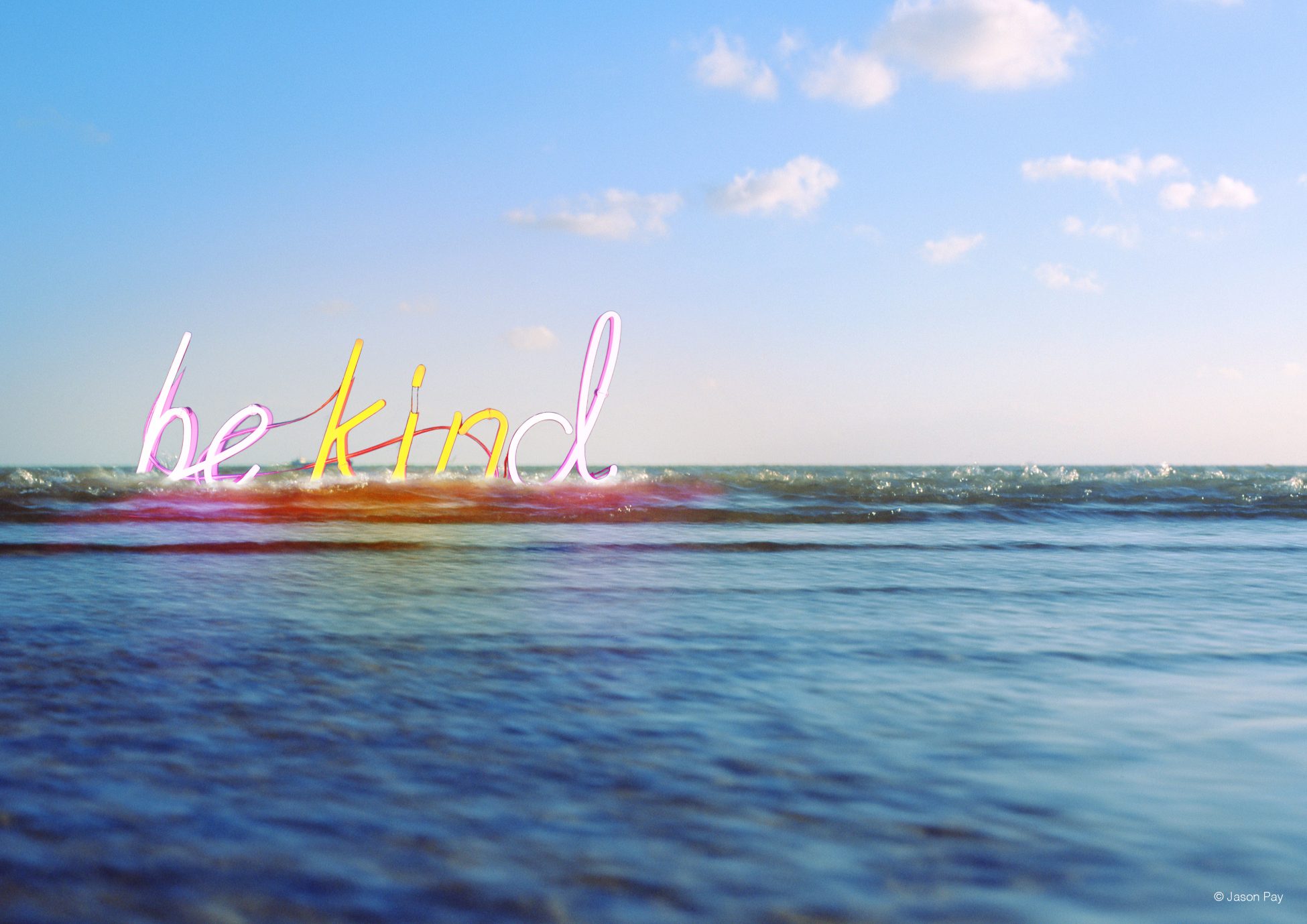 Digital artwork. A photo looking out from the shore at mostly calm seas with a small wave breaking nearby and a blue sky with a couple of small white clouds. Where the wave is breaking is a large neon sign spelling out the words 'be kind' in yellow and pink lighting.