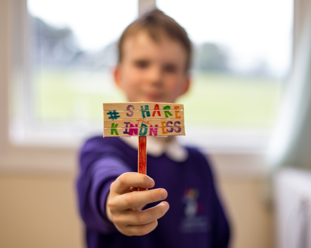 Photo of a child in a school uniform, holding a small sign made from lollipop sticks that says 