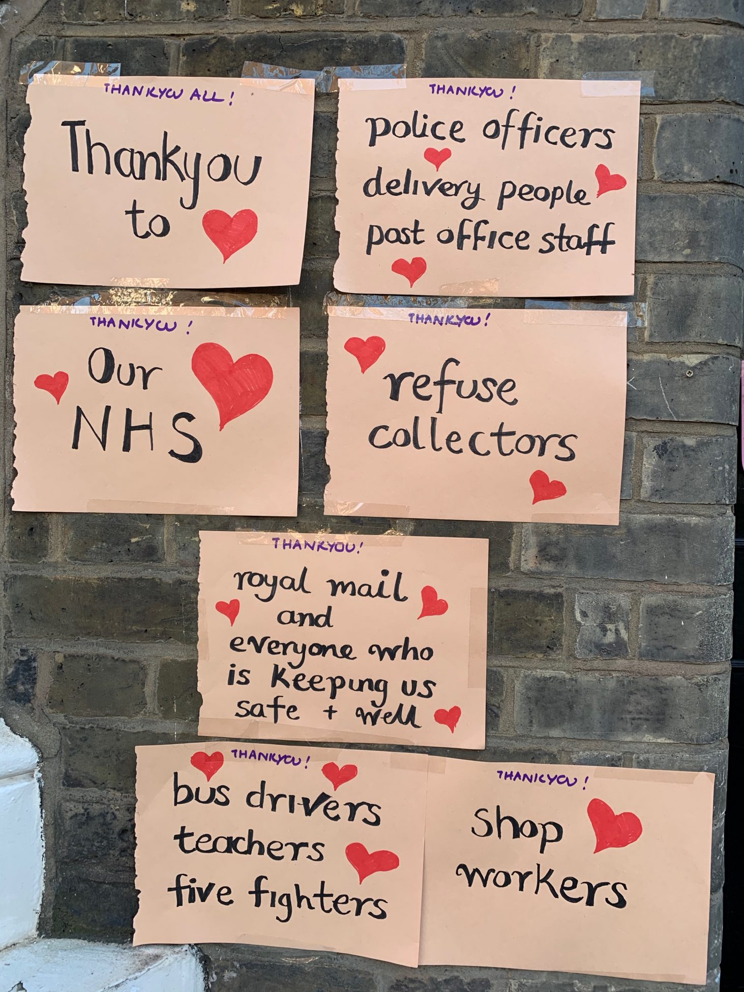 Seven hand written signs giving thanks to the NHS and key workers, decorated with red hearts and stuck up on a brick wall.