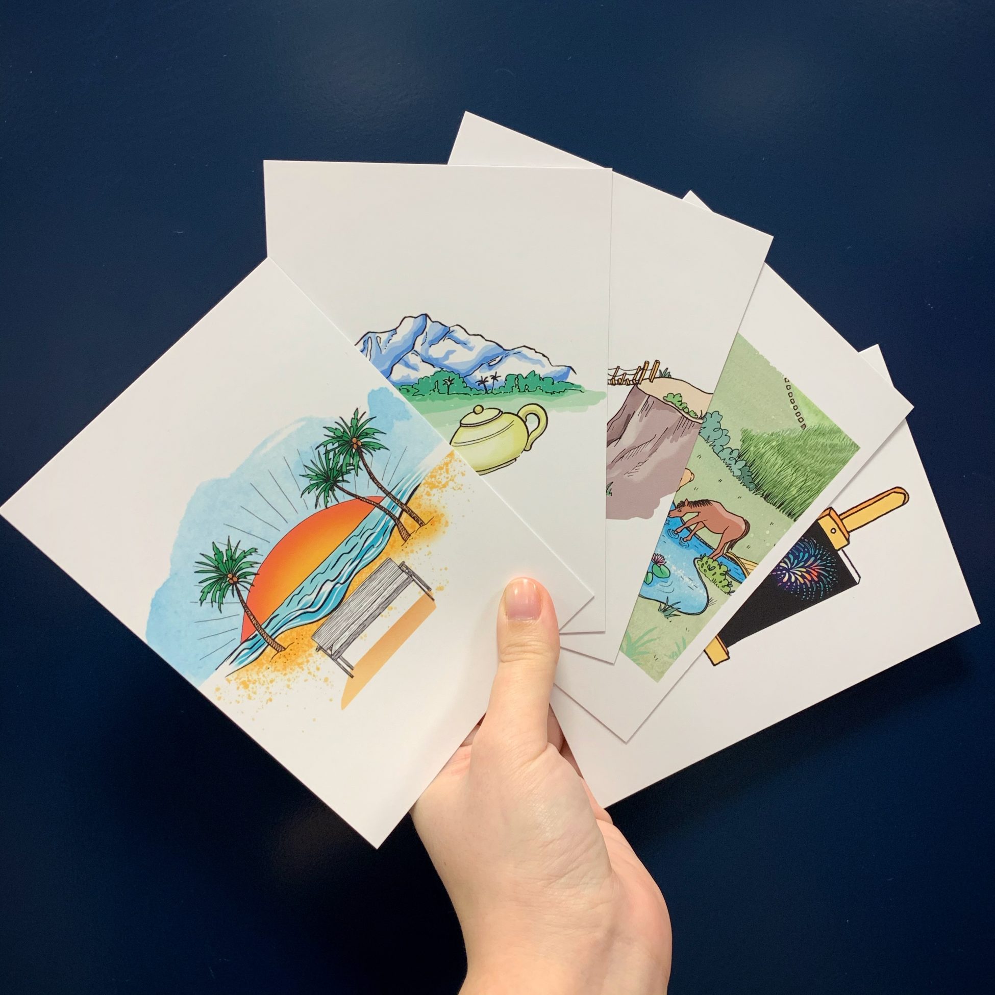 Photo of a hand holding five fanned out A6 cards, each with an illustration