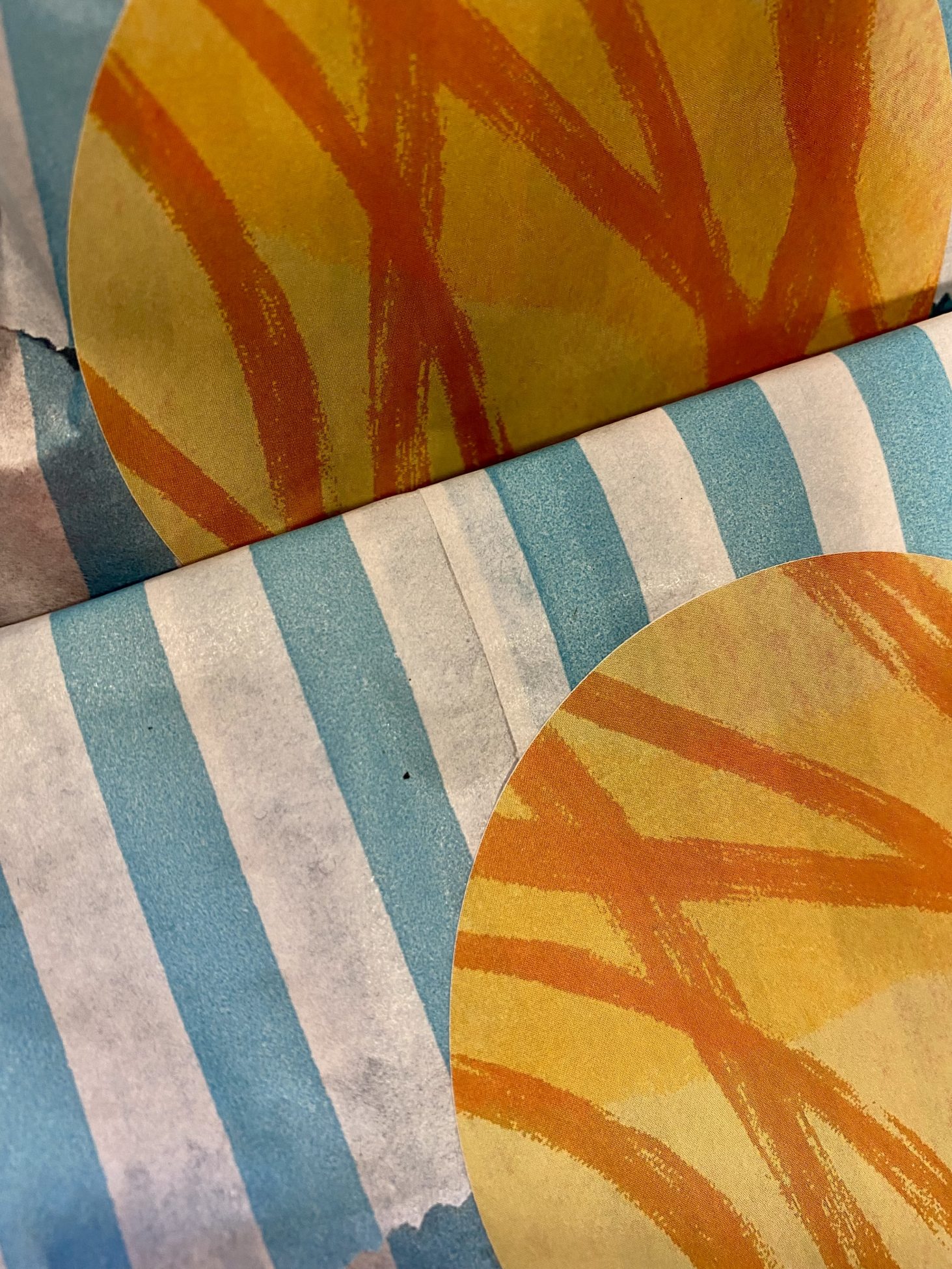 A close up photo of filled blue and white striped paper bags. The top of each bag has been folded over and sealed with a round orange sticker with abstract darker orange lines drawn across it.