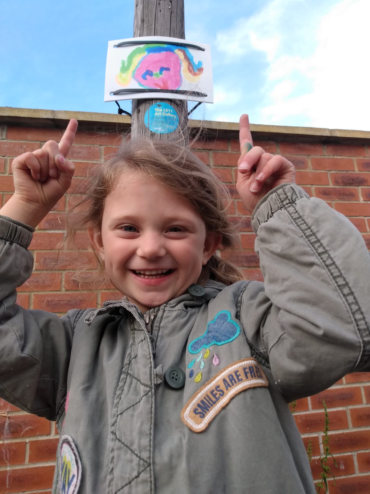 Young girl photographed in front of a lamppost, pointing with both hands up to her hand-drawn picture which has been displayed on the lamppost. She is smiling at the camera.