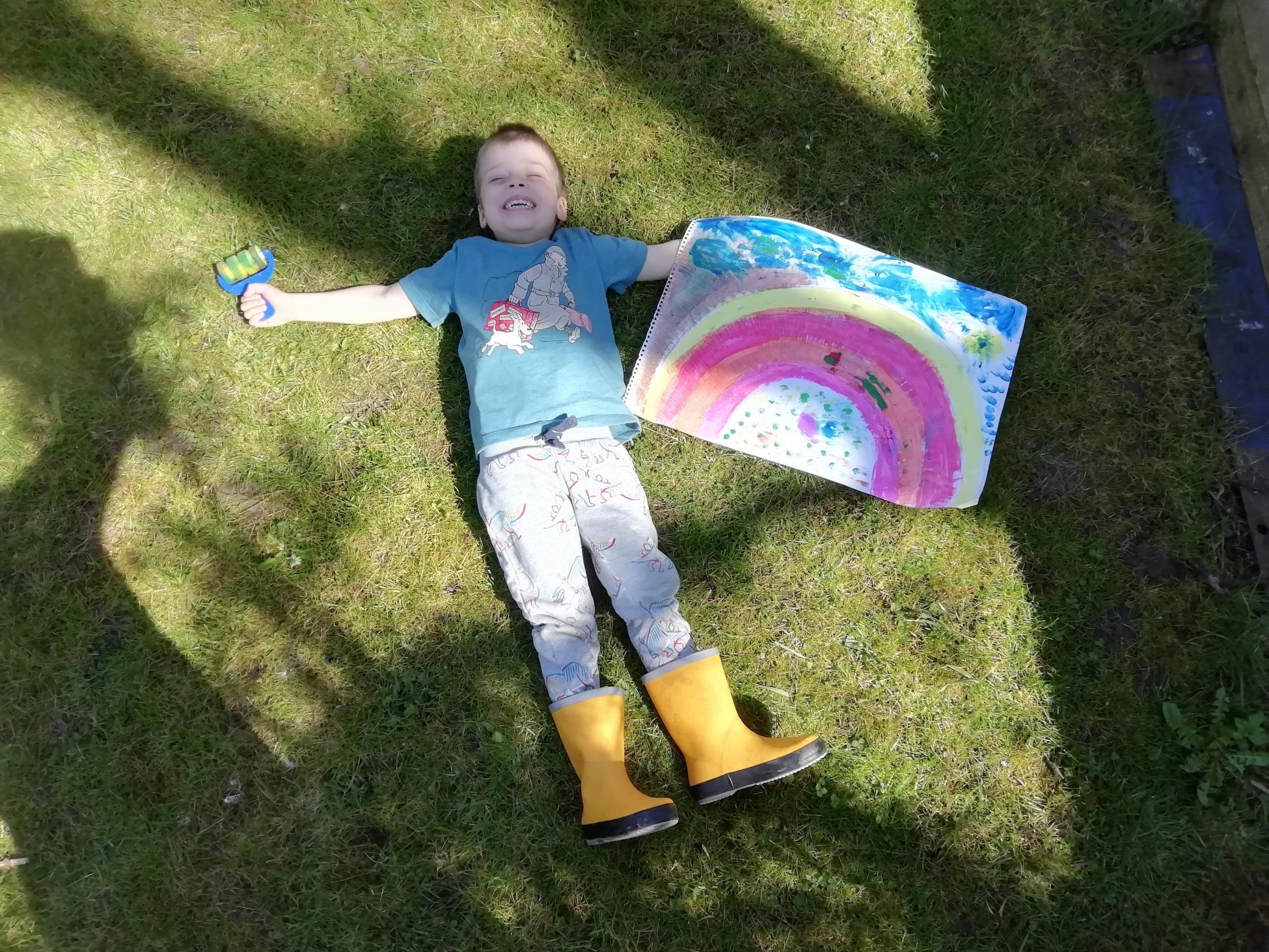 A young boy wearing blue jeans, a blue t-shirt and yellow wellies lying on the sun-lit grass next to his painting of a rainbow.