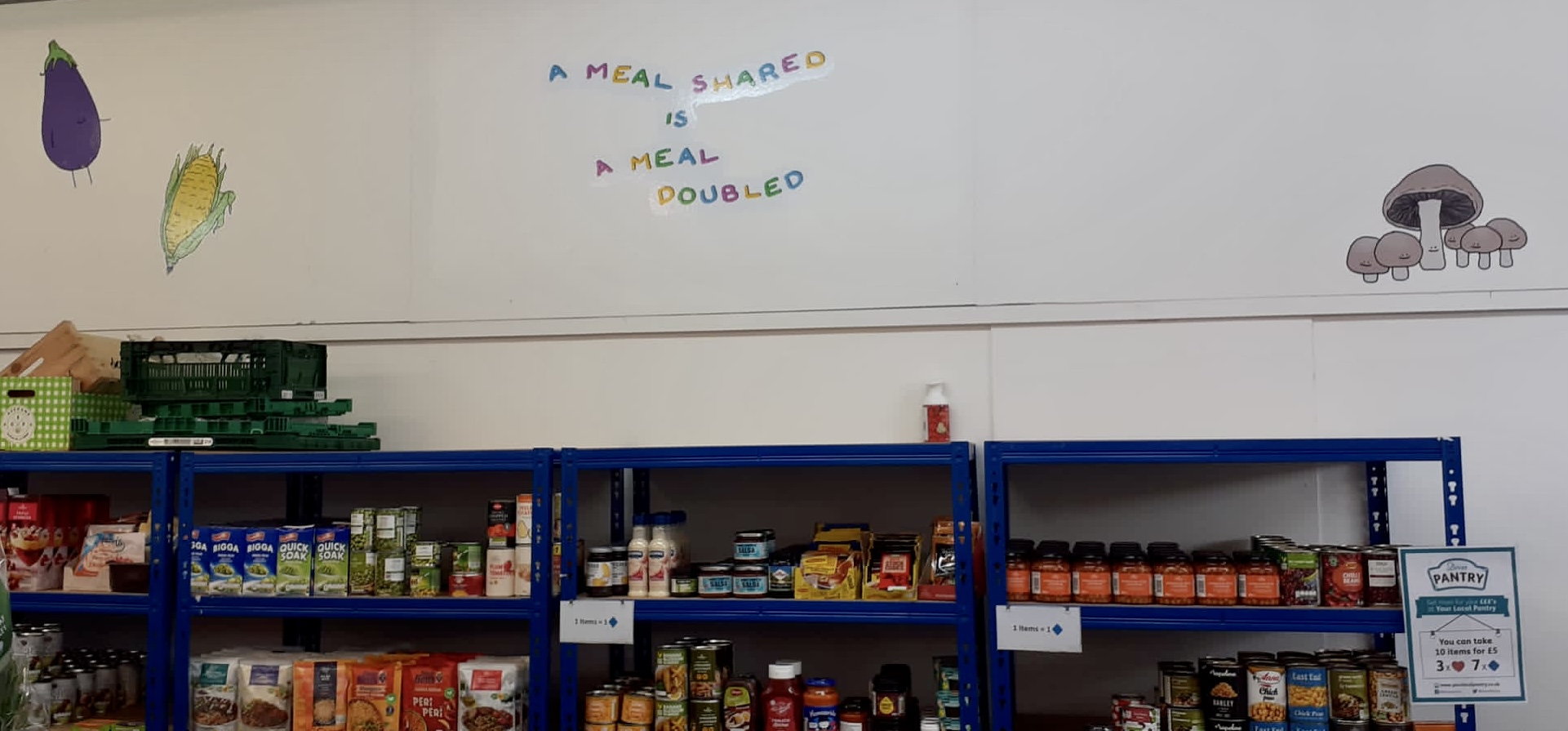 Photo of a row of metal shelving units stacked with various tins, jars and packets of food. Above the shelving are vinyl wall decals of anthropomorphised vegetables and the phrase 