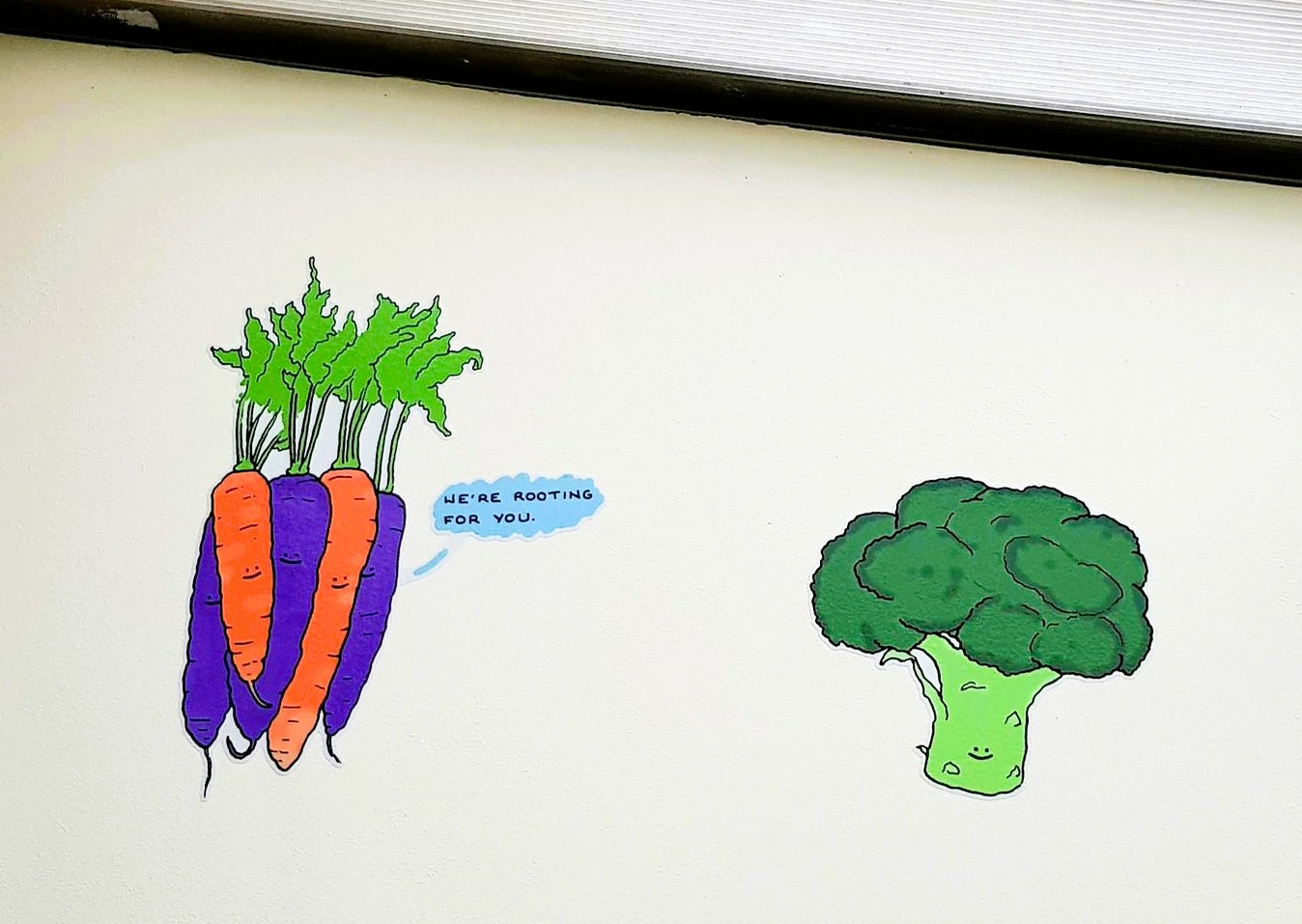 Photo of vinyl decals on a wall, they are anthropomorphised broccoli and carrots. One of the carrots is saying 