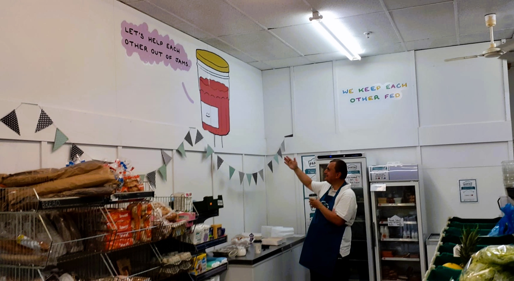 Photo of a person standing in a food pantry with racking of food and other groceries, gesturing up at a vinyl decal showing an anthropomorphised jar of jam saying 