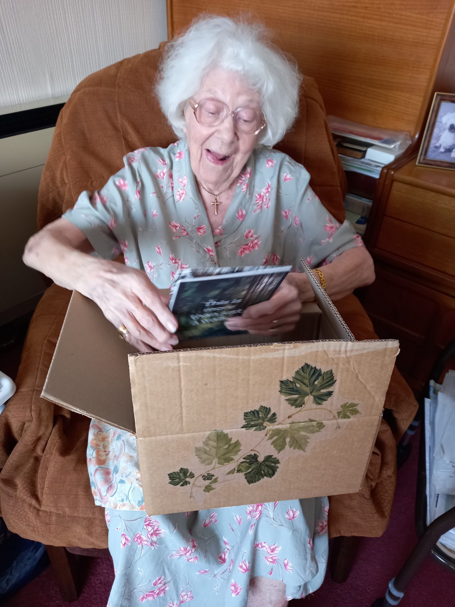 A photo of an older lady with white hair receiving and opening one of the forest bathing boxes. She is looking at the contents of the box and smiling.