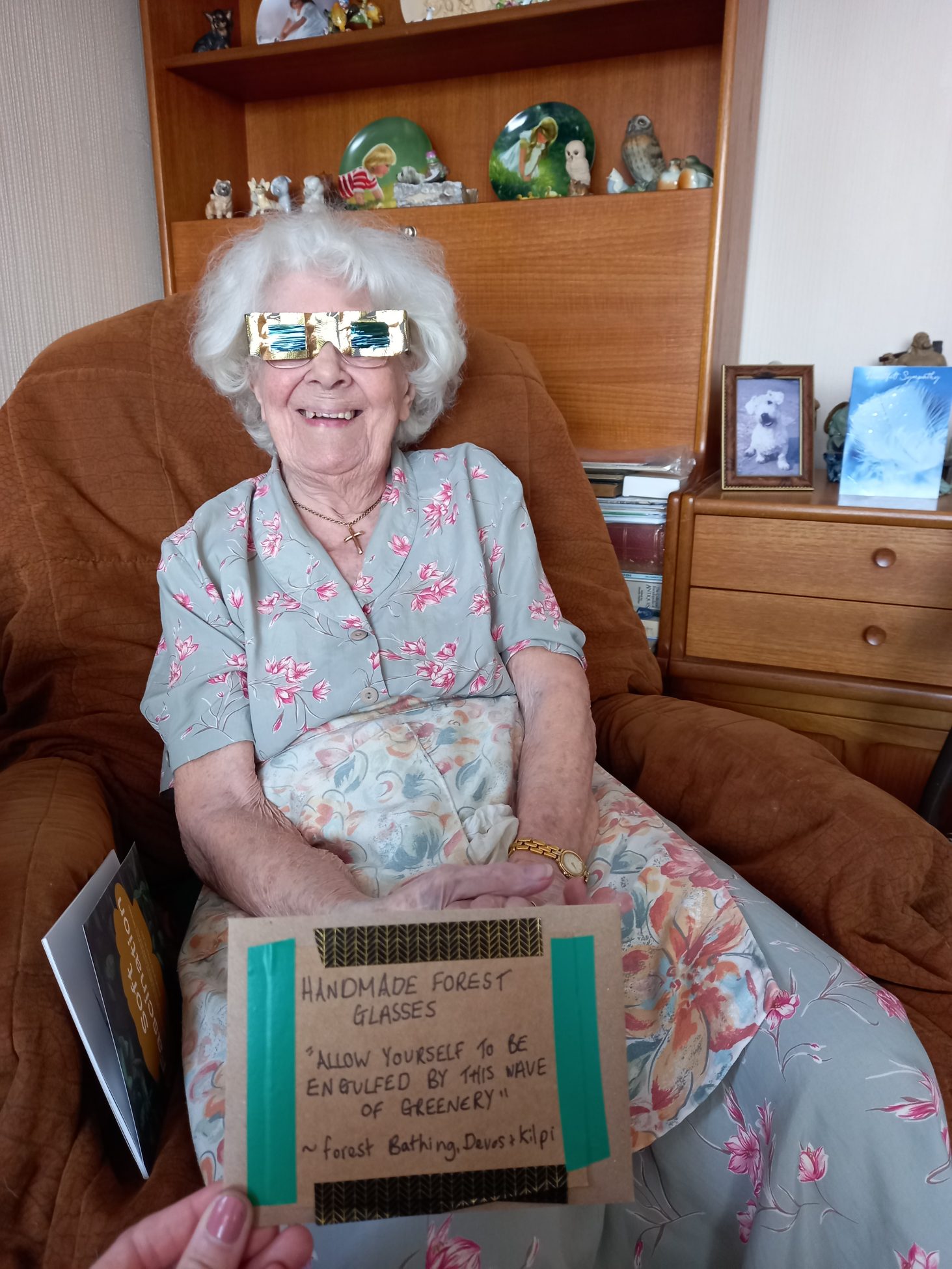 A photo of an older lady with white hair trying on the forest glasses from her forest bathing box. She is smiling.