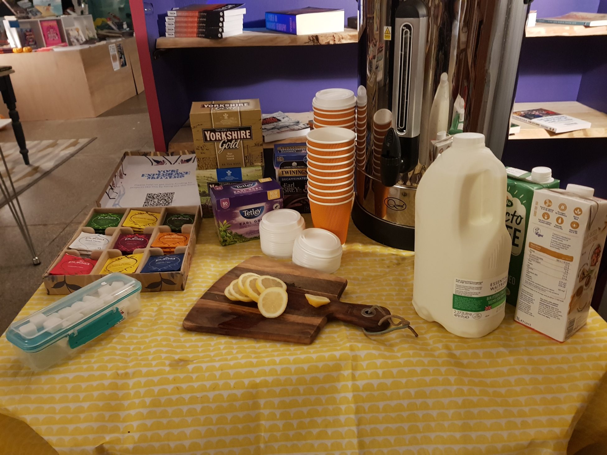 Photo of a small table in front of a shelf of books. The table has a vibrant yellow table cloth on it, and a selection of teas and herbal infusions along with a large urn, a selection of milk and non-dairy alternatives, slices of lemon, sugar cubes, and recyclable hot drink cups.