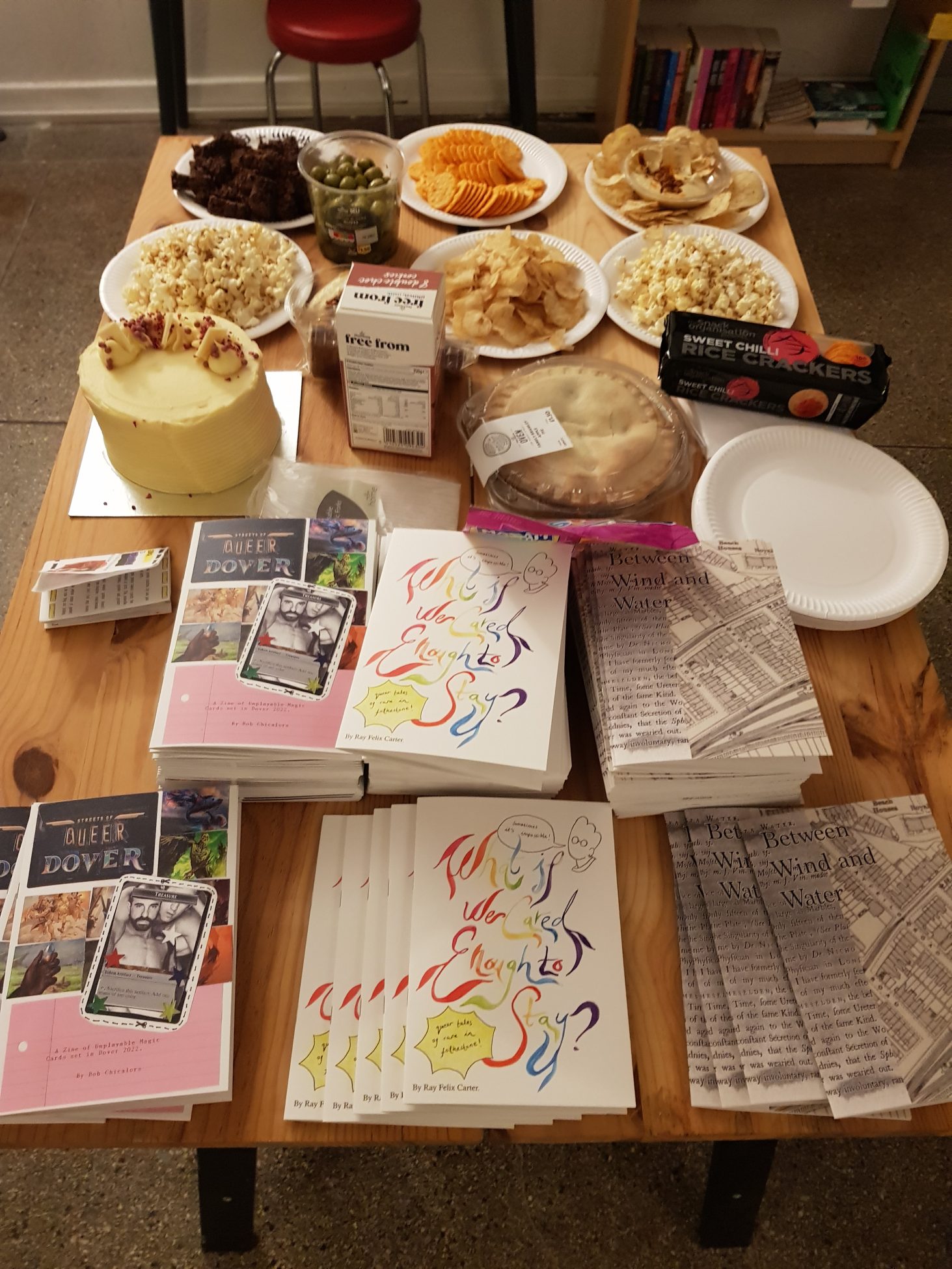 Photo of a table in Folkestone Bookshop. One half has stacks of the three zines, printed as A5 booklets, and the other half has a variety of snacks including pop corn, olives, cakes, crisps and crackers.