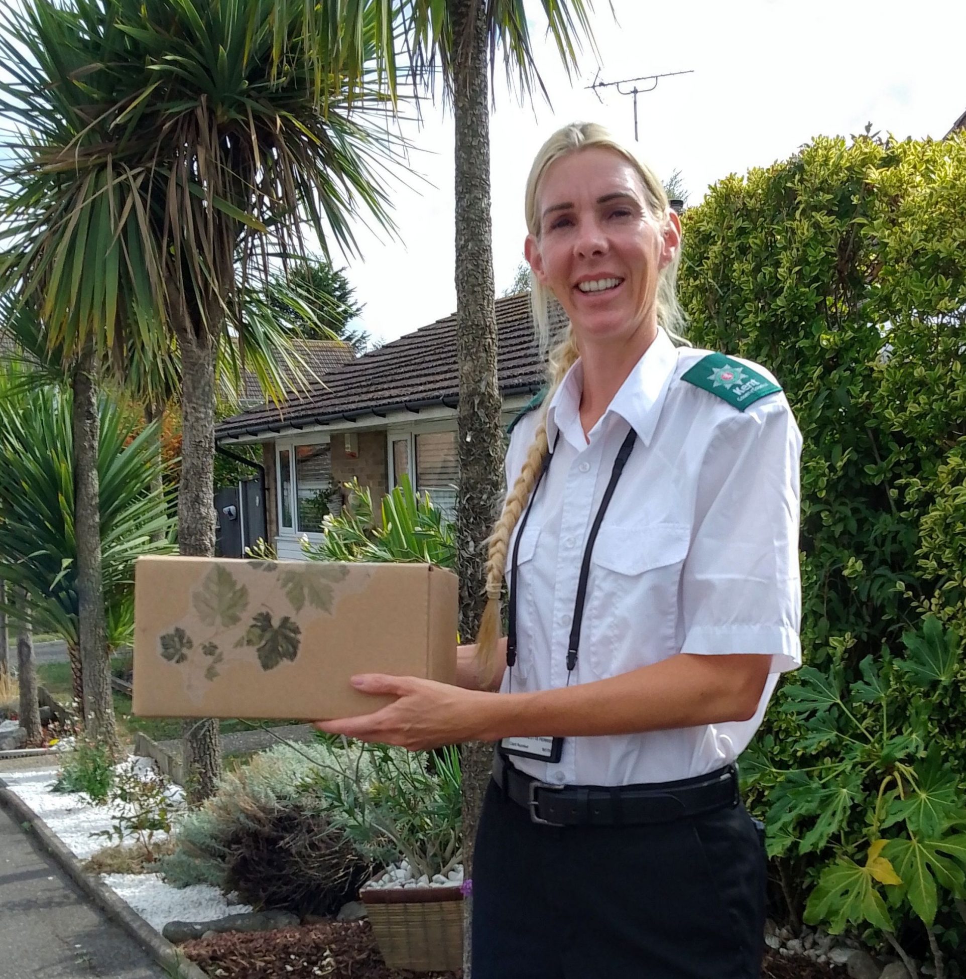A community warden delivering one of the forest bathing boxes. She is blonde with a pony tail and is wearing a white and green smart uniform.