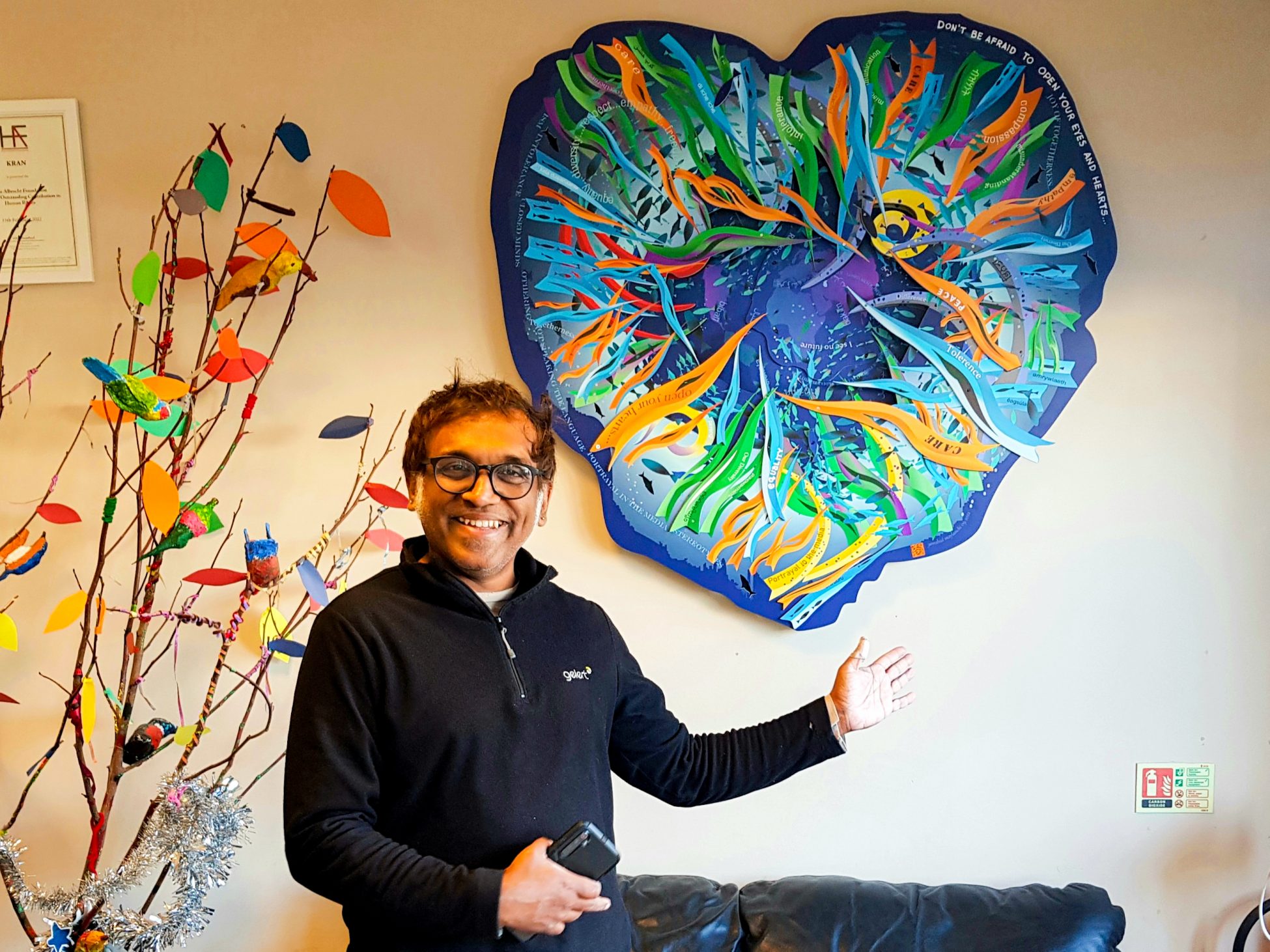 A photo of artist Jamshid Maylanchi standing in front of the heart shaped artwork and gesturing towards it whilst smiling at the camera.