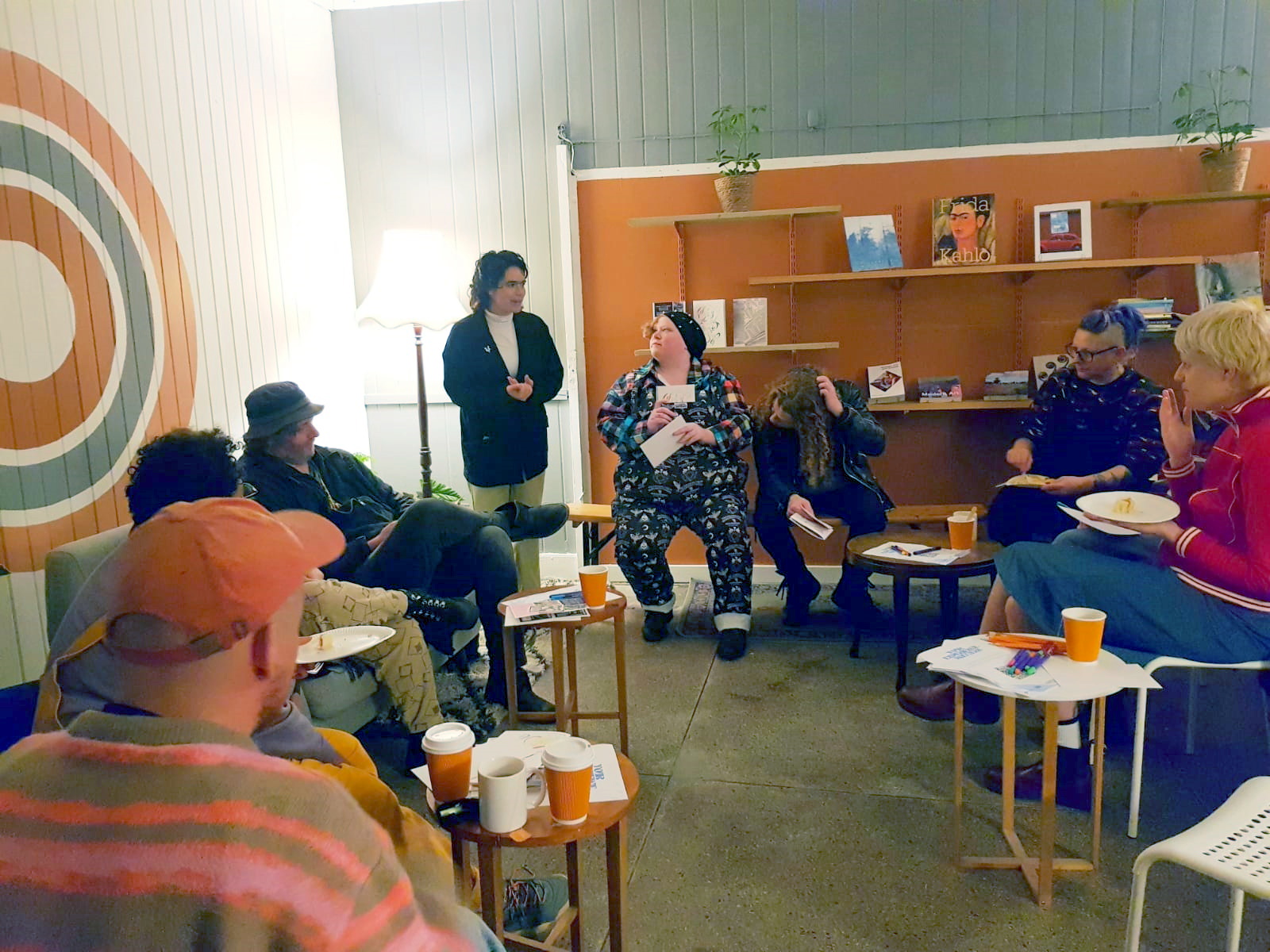 Photo of a brightly painted bookshop with various mismatched chairs and side tables. Dre Spisto is standing in one corner speaking to the attendees, who are sitting to their right, while the three zine artists are sitting to their left.